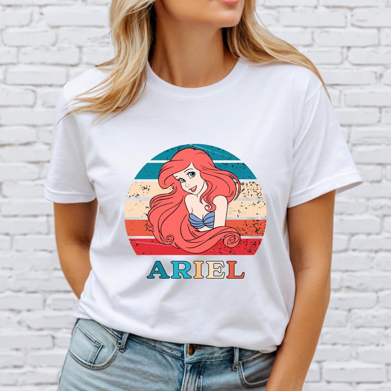 Puede incluir: Camiseta blanca con un gr&aacute;fico de estilo vintage de Ariel de La Sirenita. El gr&aacute;fico presenta a Ariel con el pelo rojo, un sujetador de concha azul, una cola roja y un fondo de puesta de sol. El texto "ARIEL" est&aacute; impreso debajo del gr&aacute;fico.