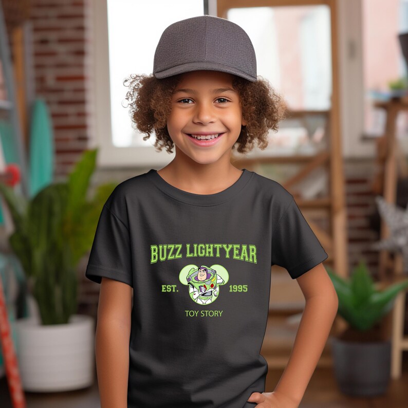 May include: A black t-shirt featuring the text "Buzz Lightyear" in green, with an image of the character and the text "Toy Story". The shirt also includes the text "Est. 1995". The child is wearing a gray baseball cap.