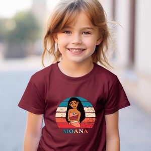 Puede incluir: Una camiseta de color granate con un gr&aacute;fico retro de Moana, una princesa polinesia de la pel&iacute;cula de Disney Moana. El gr&aacute;fico presenta una puesta de sol con un dise&ntilde;o a rayas de colores y el texto "MOANA".