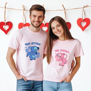 May include: Two people wearing matching pale pink t-shirts. One shirt features a blue cartoon character with the text "HER STITCH". The other shirt has a pink cartoon character and the text "HIS ANGEL". Red heart decorations hang in the background.