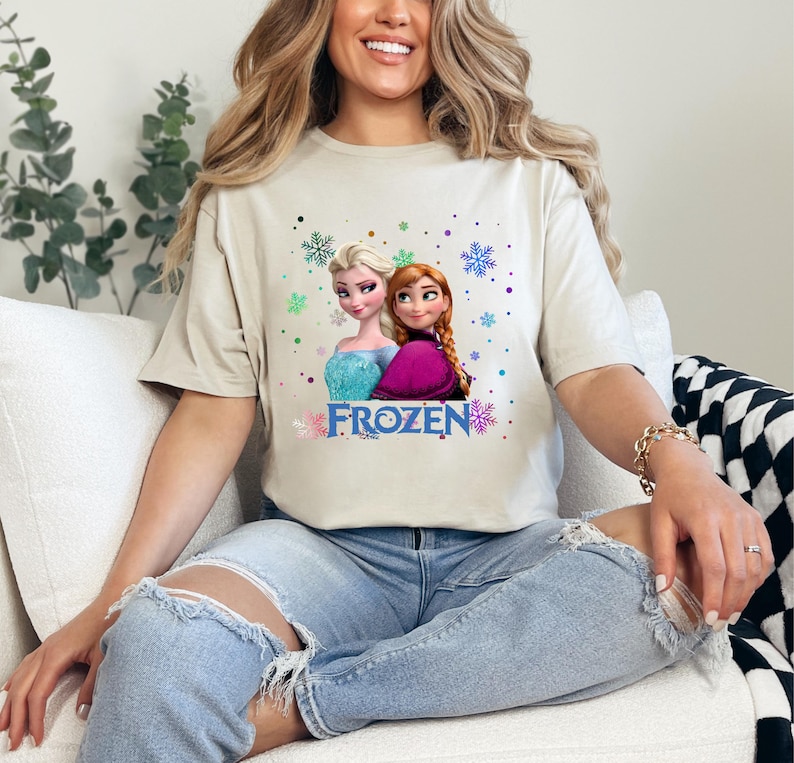 May include: A white t-shirt with a graphic of the characters Elsa and Anna from the movie Frozen. The graphic includes the word "FROZEN" and snowflakes.
