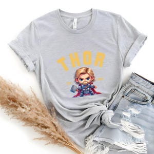 K&ouml;nnte beinhalten: Hellgraues T-Shirt mit einer Cartoon-Illustration von Thor und dem Wort "THOR" in Gelb. Die Figur tr&auml;gt einen Hammer und einen roten Umhang. Das T-Shirt wird mit hellblauen Jeansshorts kombiniert. Das T-Shirt ist aus weichem Material.