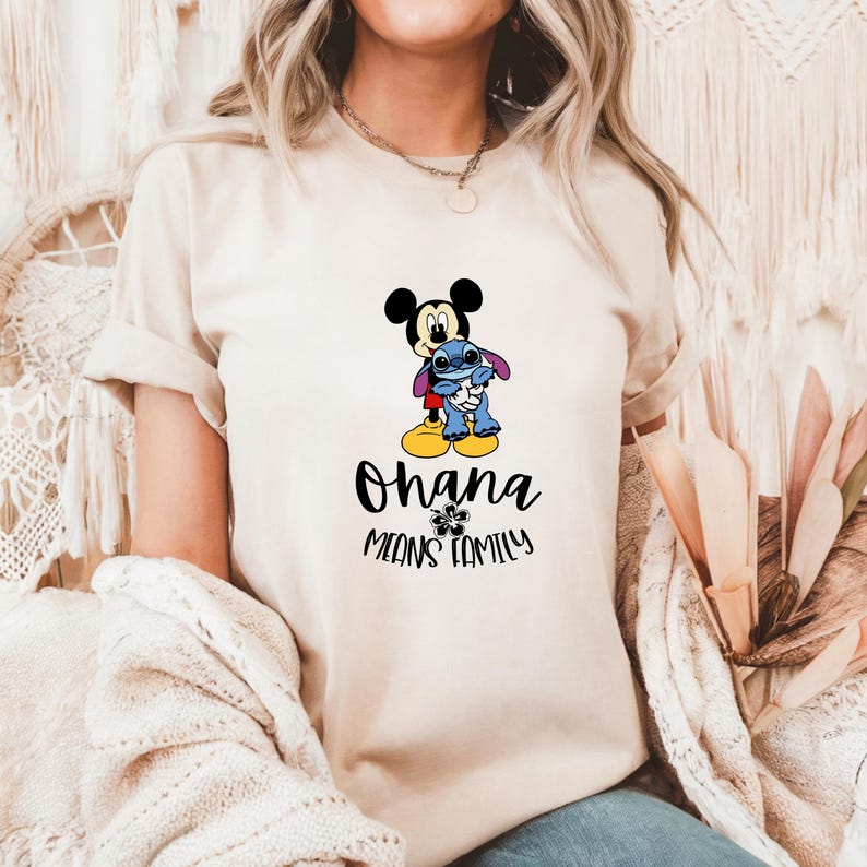 May include: Beige t-shirt featuring a graphic of Mickey Mouse hugging Stitch. Below the characters, the text reads "Ohana means family" in black script. The design includes a small flower graphic.