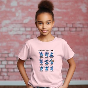 May include: A light pink t-shirt featuring a graphic of the character Stitch from Lilo & Stitch. The graphic includes the text "GOD SAYS THAT I AM..." and various positive affirmations. The shirt is worn by a young person.