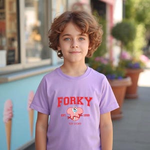 May include: A light purple t-shirt featuring the word "FORKY" in red collegiate lettering above a cartoon character illustration. The text "EST. 1999 TOY STORY" is below the illustration. The shirt is worn by a child with curly brown hair.