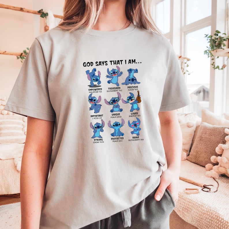 May include: A light gray t-shirt featuring the text "GOD SAYS THAT I AM..." above a grid of blue cartoon characters. Each character represents a positive affirmation, such as "Empowered" and "Loved".