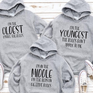 May include: Three gray hooded sweatshirts with black text: "I'M THE OLDEST I MAKE THE RULES", "I'M THE YOUNGEST THE RULES DON'T APPLY TO ME", and "I'M IN THE MIDDLE I'M THE REASON WE HAVE RULES". White sneakers are placed near the sweatshirts.