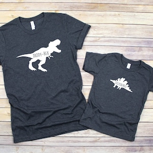 May include: Two dark gray t-shirts with white dinosaur silhouettes and text. The larger shirt says "Daddy-Rex" and the smaller shirt says "Lil'saurus".