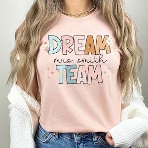 Pode incluir: Uma camiseta rosa com o texto "DREAM mrs. smith TEAM" em letras coloridas com flores e detalhes de glitter.