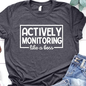 May include: Dark gray t-shirt with the text "ACTIVELY MONITORING like a boss" in a white rectangular box. The shirt is paired with blue jeans, white sneakers, and sunglasses. A sprig of greenery is also present.