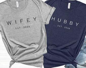May include: Two t-shirts, one grey and one navy blue, with the words "WIFEY EST 2024" and "HUBBY EST 2024" printed on them.