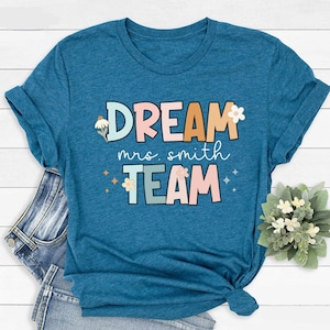 Pode incluir: Uma camiseta azul turquesa com o texto "DREAM mrs. smith TEAM" em letras coloridas com flores e brilhos.
