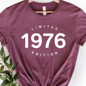 K&ouml;nnte beinhalten: Ein weinrotes T-Shirt mit hochgekrempelten &Auml;rmeln, das an einem Holzb&uuml;gel h&auml;ngt. Auf dem Shirt steht in Wei&szlig; "LIMITED 1976 EDITION". Ein Zweig mit Gr&uuml;nzeug liegt unten. Ein l&auml;ssiges Kleidungsst&uuml;ck.