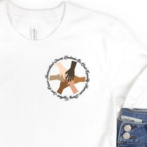 May include: A white t-shirt with a circular graphic of hands of different skin tones, with the text "Spread Love, Choose Kindness, Be Kind, Stand Together, Grow Together". The shirt is paired with denim shorts, sunglasses, and brown flip-flops.