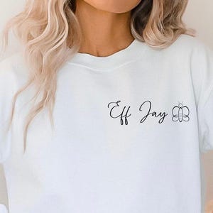 May include: White sweatshirt with long sleeves. The text "Eff Jay" is printed on the upper left chest area, accompanied by a small butterfly design. The model is wearing blue jeans.