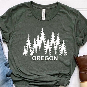 May include: A dark green t-shirt with a white graphic of a forest silhouette and the word "OREGON" underneath. The t-shirt is paired with blue jeans and brown leather boots.