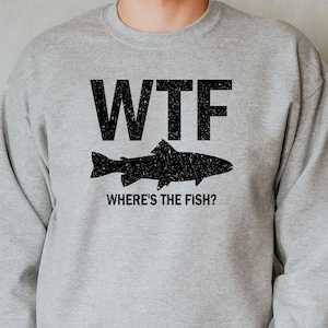 May include: A light gray sweatshirt with the text "WTF" above a black fish graphic and the phrase "WHERE'S THE FISH?" below. The sweatshirt is worn by a person in blue jeans.