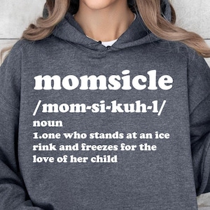 Op de afbeelding: Donkergrijze hoodie met het woord "momsicle" in witte letters. Daaronder een fonetische uitspraak en een definitie: "iemand die op een ijsbaan staat en bevriest voor de liefde van haar kind."