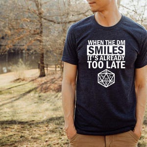 May include: A dark blue t-shirt with white text that reads "When the DM Smiles It's Already Too Late". A white 20-sided die is printed below the text.