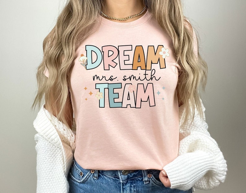 Pode incluir: Uma camiseta rosa com o texto "DREAM mrs. smith TEAM" em letras coloridas com flores e detalhes de glitter.
