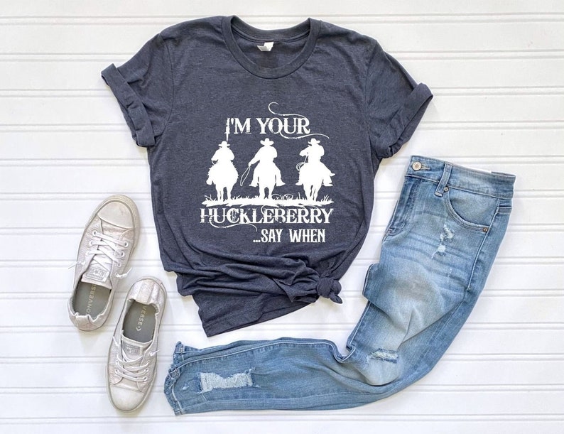 May include: A dark blue t-shirt with a white graphic of three cowboys riding horses. The text on the shirt reads "I'm your huckleberry...say when."