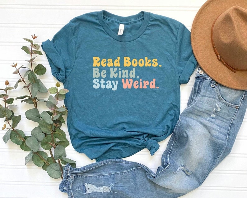 May include: Teal blue t-shirt with the text "Read Books. Be Kind. Stay Weird." in a retro font with yellow, orange, and pink colors.