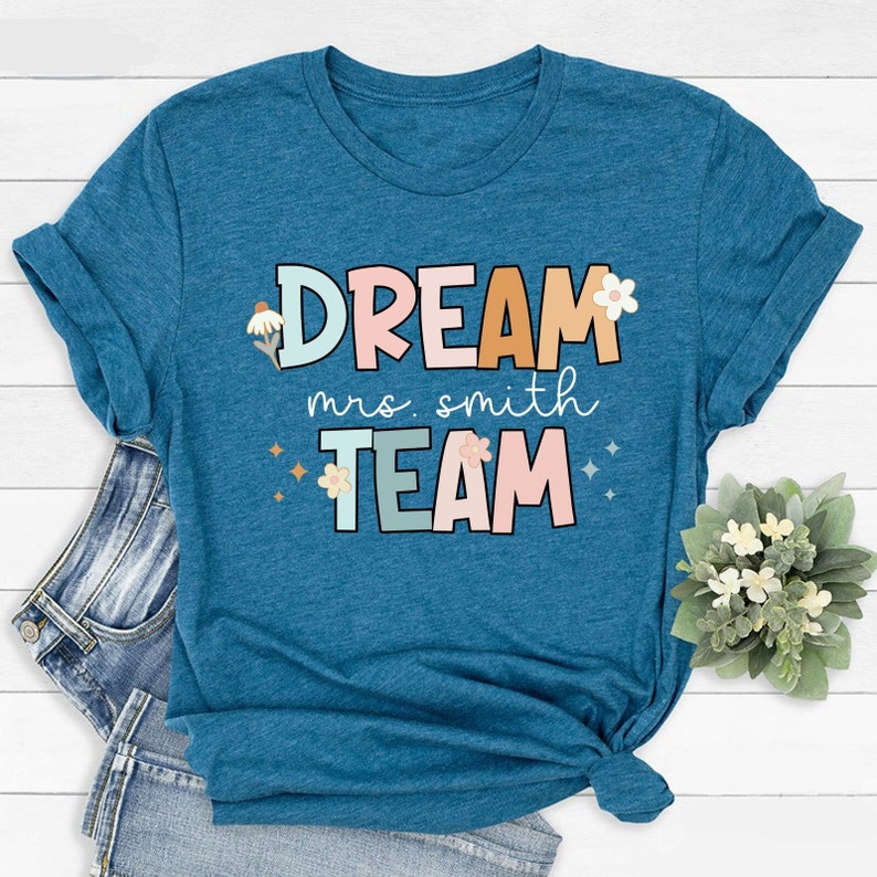 Pode incluir: Uma camiseta azul turquesa com o texto "DREAM mrs. smith TEAM" em letras coloridas com flores e brilhos.