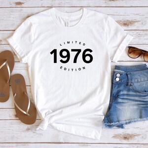 K&ouml;nnte beinhalten: Wei&szlig;es T-Shirt mit dem schwarzen Text "LIMITED 1976 EDITION". Das Shirt wird mit Jeansshorts, braunen Sandalen und Sonnenbrille kombiniert. Die Artikel sind auf einer hellen Holzmaserung angeordnet.