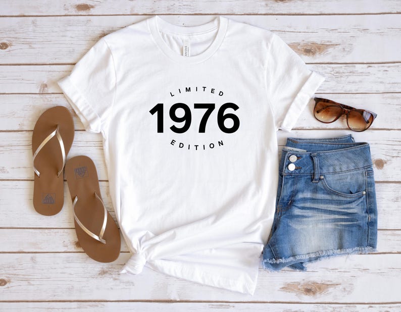 K&ouml;nnte beinhalten: Wei&szlig;es T-Shirt mit dem schwarzen Text "LIMITED 1976 EDITION". Das Shirt wird mit Jeansshorts, braunen Sandalen und Sonnenbrille kombiniert. Die Artikel sind auf einer hellen Holzmaserung angeordnet.