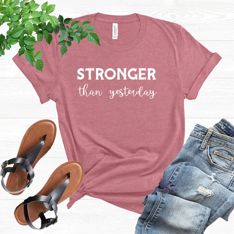 May include: A dusty rose t-shirt with the text "STRONGER than yesterday" in white. The shirt is styled with a pair of brown sandals with black straps and blue denim jeans. Green leafy vines are in the background.