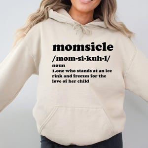 Op de afbeelding: Een beige hoodie met een voorzak en een capuchon met trekkoord. De hoodie heeft het woord "momsicle" in vette zwarte letters, samen met een fonetische uitspraak en een definitie: "iemand die op een ijsbaan staat en bevriest uit liefde voor haar kind."