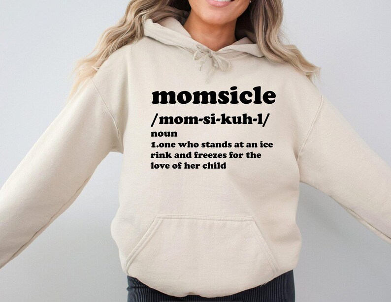 Op de afbeelding: Een beige hoodie met een voorzak en een capuchon met trekkoord. De hoodie heeft het woord "momsicle" in vette zwarte letters, samen met een fonetische uitspraak en een definitie: "iemand die op een ijsbaan staat en bevriest uit liefde voor haar kind."