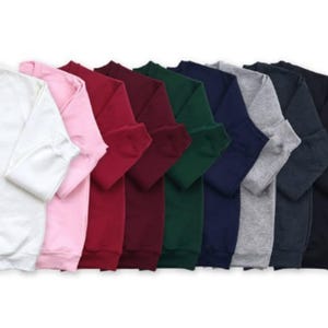Op de afbeelding: Een verzameling sweatshirts met lange mouwen en een ronde hals in verschillende kleuren: wit, roze, rood, bordeaux, groen, marineblauw, grijs en zwart. De sweatshirts zijn opgevouwen en tonen de beschikbare tinten.