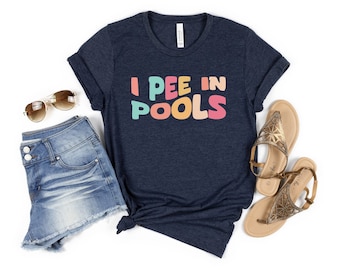 Könnte beinhalten: Ein marineblaues T-Shirt mit dem Text "I PEE IN POOLS" in Regenbogenfarben. Das Shirt wird mit Jeans-Shorts und goldenen Sandalen kombiniert.