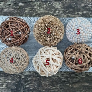 May include: Six decorative spheres made of natural materials. The spheres are arranged in a row on a grey surface. The spheres are numbered 1 through 6. Sphere 1 is brown and made of woven twigs. Sphere 2 is beige and made of woven fabric. Sphere 3 is white and made of small beads. Sphere 4 is beige and made of woven twine. Sphere 5 is beige and made of woven strips. Sphere 6 is brown and made of woven twigs.