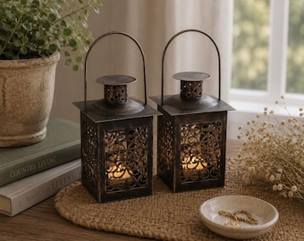Puede incluir: Dos faroles cuadrados de metal con acabado en bronce oscuro y patrones de filigrana recortados en los laterales. Cada farol cuenta con un asa superior, una chimenea cilíndrica con detalles perforados y una pequeña vela encendida dentro.