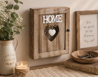 May include: A rectangular wooden key cabinet with a vertical plank design, featuring the white text "HOME" at the top and a central heart-shaped cutout. A small white heart ornament hangs from a jute bow inside the cabinet, which holds metal keys.