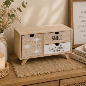 Könnte beinhalten: Ein Schreibtisch-Organizer aus hellem Holz mit drei Schubladen. Die linke Schublade hat ein weißes geometrisches Rautenmuster. Die obere rechte Schublade ist mit dem Wort SMILE beschriftet, die untere rechte trägt das Wort Everyday mit Pfeil.