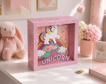 Può includere: Un salvadanaio quadrato rosa glitterato a forma di teca con una fessura sulla parte superiore. L'interno presenta un unicorno bianco su un arcobaleno colorato con nuvole e la scritta Always be a UNICORN in lettere viola su sfondo rosa.