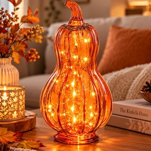 Può includere: Una zucca alta a forma di zucca ornamentale in vetro arancione traslucido, caratterizzata da dettagli a coste verticali e un picciolo curvo. L'interno è illuminato da una serie di luci bianche calde che creano un effetto luminoso.