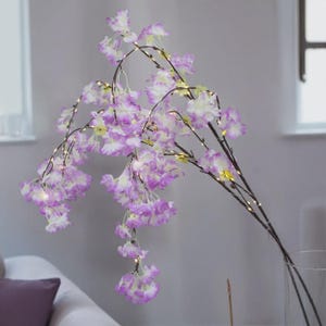 May include: Artificial floral arrangement featuring cascading branches with purple and white blossoms. The branches are adorned with small, warm white LED lights, creating a soft glow. The arrangement is displayed in a clear glass vase, adding a touch of elegance.