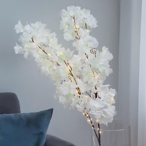 Puede incluir: Una gran rama de cerezo blanco artificial con delicados pétalos en capas. Las ramas marrones están adornadas con pequeñas luces blancas cálidas. El arreglo está en un jarrón de vidrio transparente, creando una exhibición suave y elegante.