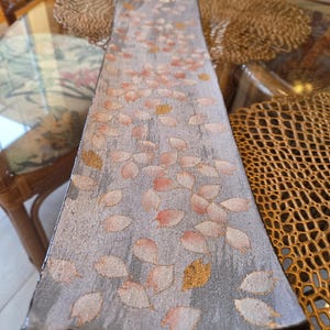 May include: A long, rectangular decorative tray with a gray background and a pattern of falling leaves in shades of pink and gold. The tray has a dark brown border and is placed on a glass table.
