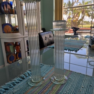 May include: Two clear glass candle holders with a ribbed column design. Each holder has a circular base and a top with a flat, circular surface. The candle holders are of different heights and sit on a blue and multicolored rug.