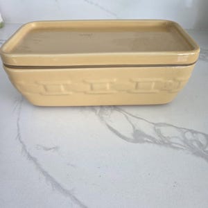 May include: A rectangular, light yellow ceramic butter dish with a matching lid. The base features a basket weave design, and the lid is flat. The dish is designed to hold butter and keep it fresh.