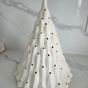 May include: A white ceramic peacock-themed Christmas tree with a textured surface. The tree features a conical shape with a pointed top and is adorned with numerous small, dark holes and feather-like details.