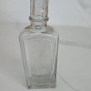 May include: A clear, antique glass bottle with a rectangular body and a detailed, cylindrical neck. The bottle has a slightly worn appearance, with visible imperfections in the glass. The design suggests it may have been used for medicine or perfume.