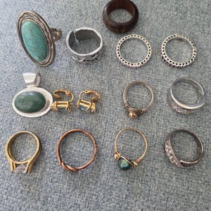 May include: A collection of jewelry including a large oval turquoise ring, a dark wood band, a silver-tone textured ring, two silver-tone eternity bands, a green stone pendant, two gold-tone clip-on earrings, a wire-wrapped ring, and several metal bands.