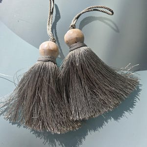 May include: Two gray decorative tassels with wooden beads and twisted cord loops. The tassels have a full, flowing design, and the beads are a light beige color. The cord loops are a matching gray color.