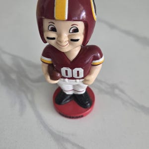 May include: A vintage figurine of a football player wearing a burgundy helmet with a yellow stripe and a matching jersey with the number "00". The figurine is holding a football and stands on a red base.